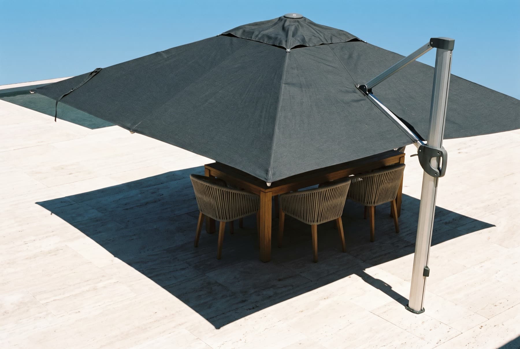 Ombrelone lateral de lona escura aberto sobre piso de pedra clara ao sol forte, com a mesa e as cadeiras inteiramente dentro da sombra projetada, imagem de referência.